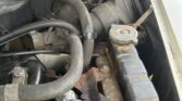 Close-up of an open MINI car hood showing the engine and various components. Focus is on black hoses, metal pipes, and a weathered radiator cap. There are signs of rust and wear on some metal parts, and wires are visible in the background. The photo was taken in daylight.