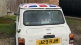 The rear view of a white MINI Cooper with a UK flag roof design is parked on a gravel driveway. The license plate reads "J29 RJB," and a "Mayfair" badge adorns the trunk. In the rear window, a sticker proclaims "I ❤ MY MINI." Residential buildings and a garage surround the car.