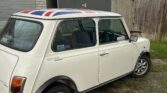 A classic white MINI Mayfair is parked on a gravel surface near a set of old, worn-out garages on a cloudy day. The car features a Union Jack roof design and silver alloy wheels. The background includes weathered wooden and metal structures, and patches of grass.
