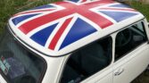 A white MINI Cooper with the British Union Jack painted on its roof is parked in front of a weathered shed and garage in an outdoor setting. The car features "Mayfair" on its side and various stickers on the rear window. The ground around the MINI is a mix of dirt, grass, and gravel.
