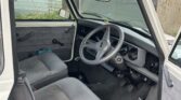 The image shows the interior of an old, white, right-hand drive MINI with cloth seats and a manual transmission. The driver's door is open, revealing a steering wheel, speedometer, and various controls. The vehicle is parked outside near a few weathered buildings.