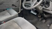 Interior of a vintage MINI car with gray upholstered seats and black flooring. The dashboard features a simple design with a speedometer and other gauges. The steering wheel is mounted on the right, indicating a right-hand drive. Visible are the gear shift, pedals, and manual window crank.
