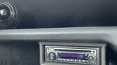A car dashboard featuring a retro-style digital car stereo mounted below the main dash area, reminiscent of MINI's classic design. The stereo has a small screen with blue digital text displaying a radio station. Surrounding the screen are various buttons and knobs, set within an interior of primarily grey and black.