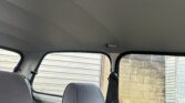 Interior view of an older MINI car featuring gray fabric seats, two in the front and a visible bench seat in the back. The seat belts are black, and the windows reveal the exterior walls of a garage with horizontal siding and a cinderblock wall. The ceiling is plain gray with a handle above the rear door.