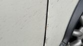Close-up of a MINI's rear left door area, showing chipped and peeling white paint with visible rust spots around the edges. The door seam is slightly misaligned, and there is a black rubber trim between the door and the wheel well. The silver alloy wheel is partially visible at the bottom right.