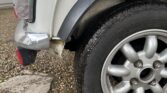 Close-up view of a silver MINI's rear wheel and bumper. The car features a silver alloy wheel with a black tire, and the body shows minor wear and some rust near the wheel arch. The ground beneath the car is covered in small pebbles. A light gray garage door is visible in the background.