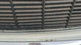 Close-up of a MINI's front grille showing vertical slats on a black background with some dirt and wear. Below the grille, part of a chrome bumper is visible, reflecting the ground and surroundings. A portion of a license plate, blacked out, can be seen at the bottom edge of the bumper.