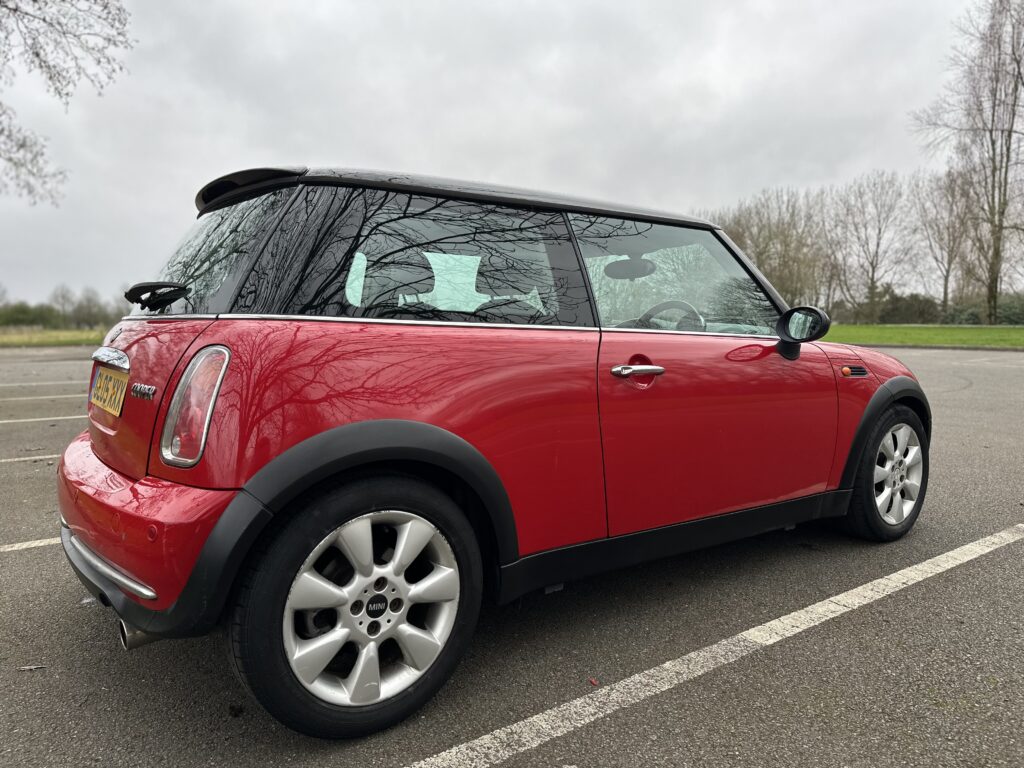 A red MINI Cooper with a black roof and side mirrors is parked in an empty outdoor parking lot on a cloudy day. The car displays a license plate on the back and has alloy wheels. Leafless trees and grass in the background reflect a late autumn or winter season.