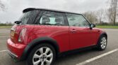A red MINI Cooper with a black roof and side mirrors is parked in an empty outdoor parking lot on a cloudy day. The car displays a license plate on the back and has alloy wheels. Leafless trees and grass in the background reflect a late autumn or winter season.