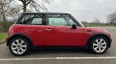 A red MINI Cooper with black roof and side mirrors is parked in an empty lot. The side view of the car highlights its compact design. Background shows overcast skies, leafless trees, and a few scattered bare branches, indicating a winter season.