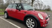 A vibrant red MINI Cooper is parked in an empty lot on a cloudy day. The car has a black roof and silver alloy wheels. Its headlights and grille are prominent. Bare trees and overcast skies form the backdrop, evoking a wintry or early spring setting for this stylish MINI.