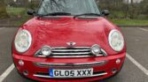Front view of a red MINI Cooper with a distinct UK license plate reading "GL05 XXX." The car is parked on a paved area with trees and a fence in the background. The vehicle features two round headlights and a pair of additional lights below them, with the MINI logo on the hood.