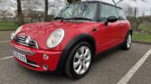 A classic red MINI Cooper with dual black racing stripes parked in an outdoor space on a cloudy day under bare trees. The car has a white number plate "XX05 XXX," fog lights, and silver alloy wheels. The background includes a fence, grassy area, and partially visible houses.