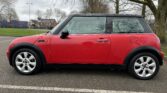 A red MINI Cooper with a black roof is parked on the side of the road. Seen from the left side, it showcases its distinctive compact design. Trees and buildings are visible in the background on a cloudy day. The car has silver alloy wheels and a side mirror with an integrated indicator light.