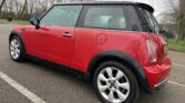 A red MINI Cooper is parked in an outdoor parking space. The car is viewed from the rear-right angle, showcasing its compact design and distinctive black roof. The rear license plate is visible, and the background features leafless trees and overcast skies typical of winter.