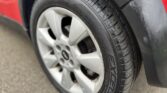 Close-up of the rear tire of a red MINI vehicle. The tire features the brand name "SYRON" and is mounted on a silver alloy rim. The surrounding black wheel arch is slightly wet, indicating recent exposure to rain or water, matching the damp, textured asphalt ground beneath the tire.