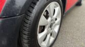 Close-up image of a MINI's front tire and alloy wheel. The tire appears to be in good condition, with visible tread patterns, and is mounted on a silver alloy rim. The car, partially visible, has a red body and is parked on a grey asphalt surface.