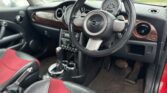 The interior of the car features a black steering wheel with a MINI logo at the center. The dashboard has a circular speedometer and wood finish accents. The seats are black and red two-toned leather. A gear shift is visible between the front seats, and the floor is carpeted in beige.