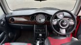 The image shows the interior of a car, specifically a MINI Cooper. It features a black dashboard with a central dial displaying the speedometer, a wooden panel, and a classic MINI Cooper logo on the steering wheel. The seats are a mix of black and red upholstery. The car is parked outdoors.