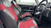 Interior of a compact MINI car featuring black and red leather seats. The steering wheel, emblazoned with the logo, is on the right side, indicating a right-hand drive vehicle. The dashboard has various controls, and a key with a fob is in the ignition. The windows display a blurred outdoor backdrop.