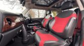 The image shows the interior of a MINI with black and red seats, a black dashboard with wooden accents, and a panoramic sunroof. The car has a right-hand drive configuration, and the windows reveal a view of trees without leaves, indicating a winter or early spring setting.