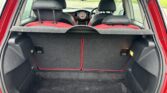 A view of the trunk of a compact MINI with the rear hatch open. The interior features a large black carpeted cargo area, flanked by dark side panels with mounted light caps and small storage pockets. The backrests of the rear seats are visible, upholstered in black and red fabric.