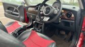 Interior view of a MINI car showcasing the driver's seat and dashboard. The car features a black steering wheel and digital speedometer. The seats are upholstered in a combination of red and black leather. The dashboard includes wood paneling and chrome accents. The foot pedals are visible below.