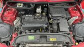 A detailed view of the engine compartment of a red Mini Cooper with the hood open. The MINI logo is prominently visible on the engine cover. Various components, hoses, and wires are neatly arranged. The surrounding area shows some dirt and water droplets, indicating recent exposure to rain or cleaning.

