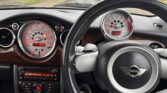 The interior of a MINI car is shown, focused on the dashboard and steering wheel. The dashboard features a large, round speedometer and a smaller tachometer next to it. The steering wheel proudly displays the MINI logo in the center. The car’s controls and radio are visible below the dashboard.