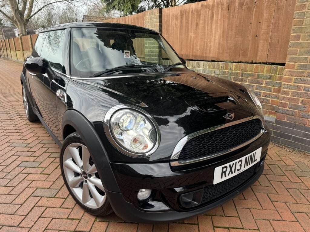 A black MINI Cooper with silver alloy wheels is parked on a brick driveway. The license plate reads "RX13 NNL." The car's exterior is shiny and clean, reflecting its surroundings. A wooden fence and part of a brick wall are visible in the background.