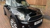 A black MINI Cooper with silver alloy wheels is parked on a brick driveway. The license plate reads "RX13 NNL." The car's exterior is shiny and clean, reflecting its surroundings. A wooden fence and part of a brick wall are visible in the background.