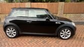 A black two-door MINI Cooper is parked on a brick driveway in front of a brick wall and wooden fence. The car, viewed from the side, has silver alloy wheels, a sloping roof, and a compact, sleek design. Tall evergreen trees stand behind the fence, providing a green backdrop.