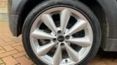 Close-up of a silver alloy wheel with a black tire mounted on a MINI car. The wheel features the iconic MINI logo at its center and is designed with multiple spokes. The tire, branded Yokohama, rests on interlocking bricks, with part of a brick wall visible in the background.