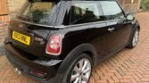 A black MINI Cooper SD with license plate "RX13 NNL" is parked on a brick driveway near a brick wall and a wooden fence. The car is viewed from the rear side, showcasing its compact design, silver alloy wheels, and distinct rear lights. Trees are visible in the background.