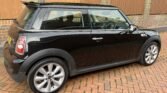 A black MINI Cooper with silver alloy wheels is parked on a herringbone-patterned brick driveway. The car is positioned facing right, in front of a brick wall topped with wooden fencing. The rear windshield wiper and compact side mirrors are visible, as well as part of the green garden in the background.