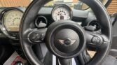 A view from the driver's seat of a MINI Cooper, showing the steering wheel with the iconic MINI emblem, central speedometer reading 0, and two smaller side gauges. The black-finished dashboard features air vents, control buttons, gear shifter, and a house and fence visible through the windshield.