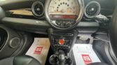 Interior of a MINI car showing the dashboard and center console. The dashboard has a large circular speedometer in the center with fuel gauge and other indicators. Below it is a small display screen with control buttons around it, and further down is the gear shift. Two AutoGlym brochures are placed on the floor mats.