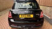 A black MINI Cooper SD is parked on a red-brick driveway nestled between two brick walls. Sporting a UK registration plate "RX13 NNL," the car features dual exhaust pipes at the bottom center of the rear bumper. The scene is serene with no other vehicles or objects in sight.