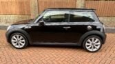 Side view of a black MINI Cooper parked on a red brick driveway. The car features silver rims, a single door on the visible side, and a silver gas cap. The background includes a wooden fence atop a brick wall, with trees and greenery visible behind the fence.