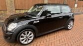 A black MINI Cooper with silver alloy wheels is parked on a red brick driveway in front of a tall wooden fence and a brick wall. The car boasts a white roof and side mirrors, complementing the surrounding greenery.