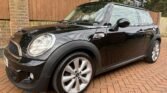 A black MINI Cooper is parked on a brick driveway. The car features silver alloy wheels, a compact two-door body, and a white roof. The background includes a wooden fence, brick wall, and dense greenery. The vehicle appears clean and polished, with a modern, sleek design.