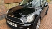 A black MINI Cooper with a UK registration plate "RX13 NNL" is parked on a brick driveway. The car boasts silver alloy wheels, a shiny exterior, and is positioned at an angle facing the camera. Brick fencing and a house are visible in the background.