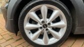 Close-up of a MINI car's silver alloy wheel featuring five pairs of spokes, branded with the MINI logo at the center. The wheel is fitted with a tire marked "205/45ZR17 88W" along the sidewall. The background shows a wet brick driveway and part of the car's grey body.