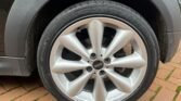 Close-up of a car tire and MINI alloy wheel on a brick-paved surface. The five-spoke wheel proudly displays the 'MINI' logo in its center. The tire's sidewall shows detailed specifications and branding information. The car's body is partially visible above the wheel.