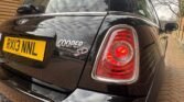 A close-up view of the rear of a black MINI Cooper SD car parked on a brick driveway. The image captures the car's tail light, rear badge reading "COOPER SD," and the yellow rear license plate with the registration "RX13 NNL." The background shows a wooden fence and some trees.