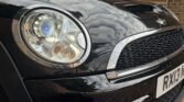 Close-up of the front left side of a black MINI Cooper. The image captures its shiny headlight with multiple bulbs, part of the grille, and hood scoop. Reflections of clouds can be seen on the car's surface. A license plate with "RX13" is partially visible at the bottom right.