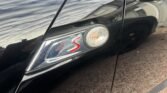 Close-up of a black MINI's side with a red "S" badge and a small circular orange indicator light on the door. The car is reflecting the sky, nearby trees, and a building. The reflection on the car's polished surface shows a distorted view of the surroundings.