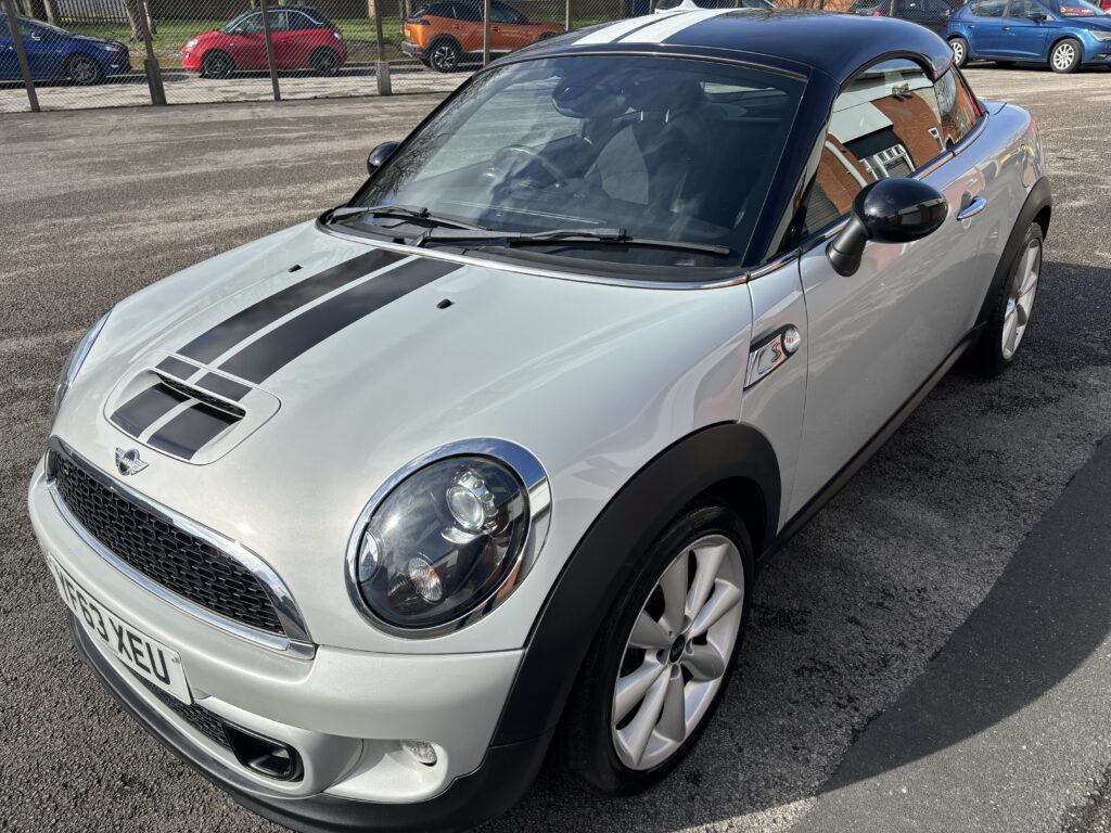 A silver MINI Cooper S with a black roof, black stripes on the hood, and white alloy wheels is parked on a paved lot. The MINI has a sporty appearance with distinctive circular headlights and a visible license plate. In the background, there is a fenced area and several other parked cars.