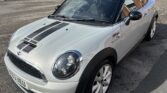 A silver MINI Cooper S with a black roof, black stripes on the hood, and white alloy wheels is parked on a paved lot. The MINI has a sporty appearance with distinctive circular headlights and a visible license plate. In the background, there is a fenced area and several other parked cars.