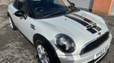 A white MINI Cooper car parked in front of a brick building. The MINI features black racing stripes on the hood and roof, a black roof, and black side mirrors. The license plate reads "YF63 XSF." The car is angled to the right with the front and driver’s side visible.